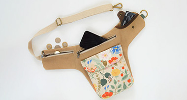 a festival hip bag on a white surface with a phone, pound coins, and a debit card sticking out of the three pockets